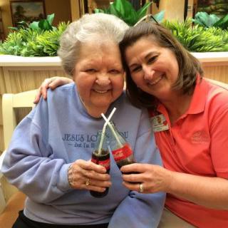 An elderly woman and her caregiver enjoy a moment together, sharing a smile and clinking Coke bottles.