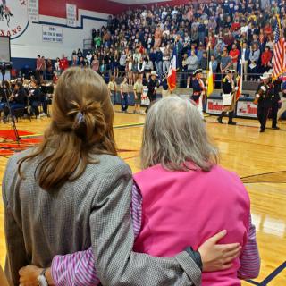 Two women embrace as a crowd gathers for a ceremony with flags in an auditorium.