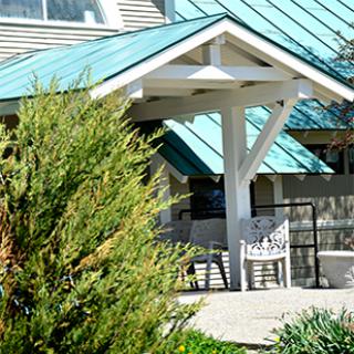 A quaint outdoor area with chairs under a sloped blue-green roof, surrounded by greenery.