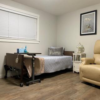 A clean, minimalist bedroom features a hospital bed with an overbed table, a recliner chair, and a side table under a window.