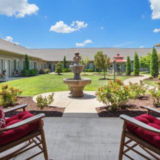 A sunny courtyard featuring a central fountain, surrounded by a neatly manicured garden and two red-cushioned chairs.
