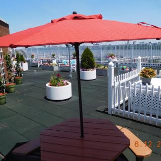 A rooftop patio with a red umbrella, surrounded by potted plants and a white fence overlooking the city.
