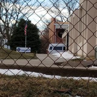A view through a chain-link fence showing a building, some snow on the ground, and a vehicle parked nearby.
