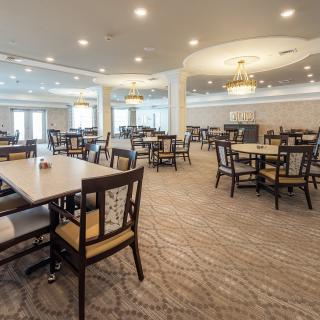 A spacious and well-lit dining hall with arranged tables and chairs, featuring chandeliers and a carpeted floor.