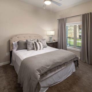 A cozy bedroom features a neatly made bed with neutral tones, complemented by a bedside lamp and window with curtains.