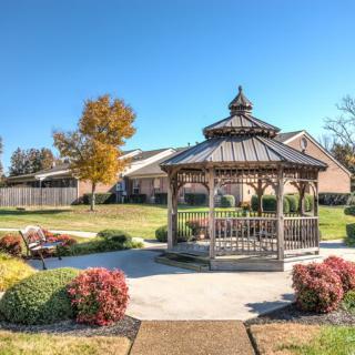 A peaceful park scene featuring a wooden gazebo surrounded by lush landscaping and a sunny sky.