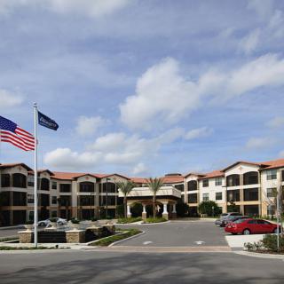 A large building with American and company flags, featuring a parking lot and landscaping under a cloudy sky.
