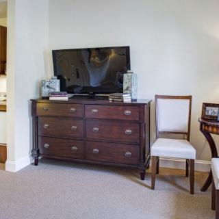 A cozy living room setup with a wooden dresser topped by a flat-screen TV and flanked by decorative items, alongside a classic chair.