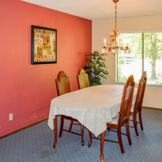 A cozy dining room with a table and four chairs, featuring a chandelier, artwork on a pink wall, and a window allowing natural light.