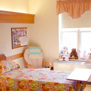 A hospital room with floral bedding, a chair, and stuffed animals by the window.
