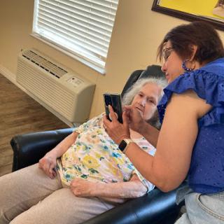 A caregiver demonstrates how to use a smartphone to an elderly woman in a cozy indoor setting.