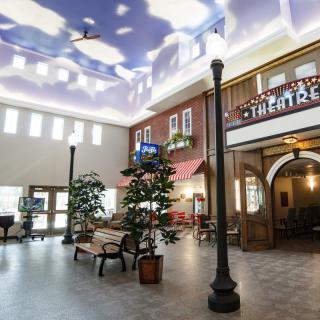 A spacious indoor atrium featuring faux storefronts, a cloud-painted ceiling, and decorative elements such as trees and lampposts.