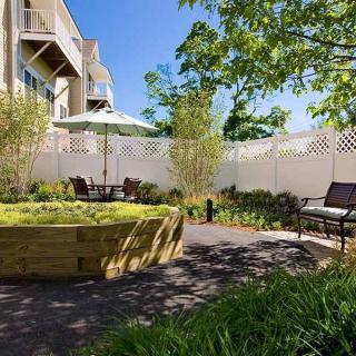 A serene outdoor garden area with seating and greenery, enclosed by a white fence next to an apartment building.