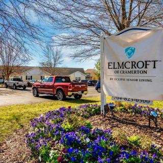 An entrance sign for Elmcroft at Cramerton with a parking area and blooming flowers.