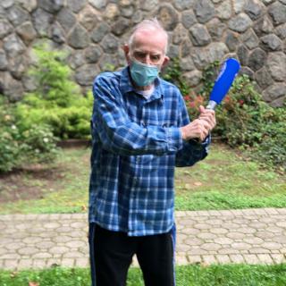 An elderly man wearing a mask stands outdoors, holding a blue object and posing in front of a stone wall.