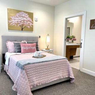 A cozy bedroom with pastel decor, featuring a bed adorned with pink and gray bedding, a side table with a lamp, and artwork of a cherry blossom tree on the wall.