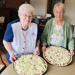 Two women proudly display their homemade pizzas, showcasing their culinary skills and joy.
