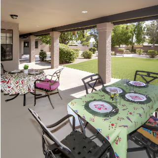 A well-arranged outdoor patio with tables and chairs under a covered porch, overlooking a green lawn on a sunny day.