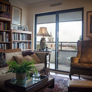 A cozy living room with a sofa, armchair, and a bookshelf, featuring a lamp and plant on the coffee table near a window leading to a balcony.