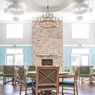 A spacious and well-lit dining area with a central stone fireplace, elegant chandelier, and neatly arranged tables and chairs.