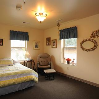 A cozy bedroom with a bed, chair, and wall decorations, softly lit by a ceiling light.