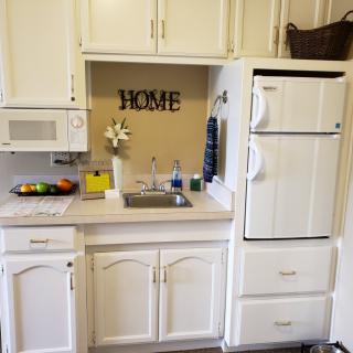 A cozy kitchen area featuring white cabinets, a sink, a refrigerator, and a microwave, with a decorative 'HOME' sign on the wall.