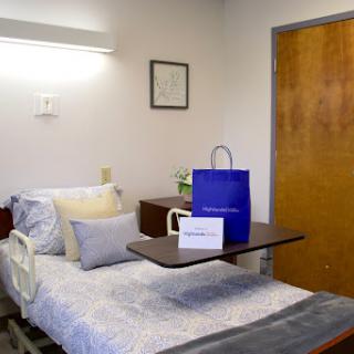 A cozy hospital room featuring a neatly made bed with decorative pillows, a small table displaying a gift bag, and warm lighting.