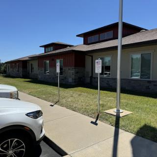 A view of a modern building with a parking area, surrounded by green grass under clear blue skies.