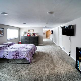 A spacious bedroom with carpeted flooring, featuring a bed with a purple comforter, a dresser, and a mounted television.