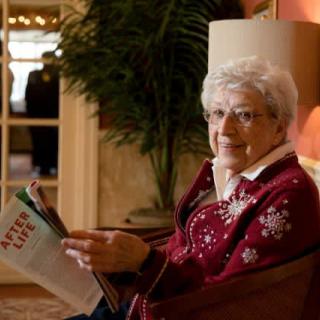 An elderly woman wearing glasses and a sweater is seated indoors, reading a magazine.