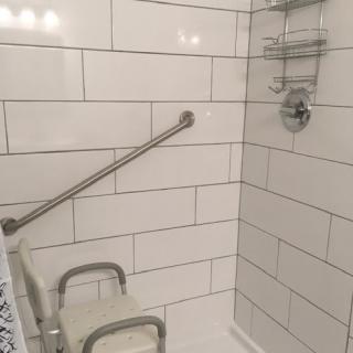 A bathroom shower with a tiled wall, equipped with a grab bar and shower seat for safety.