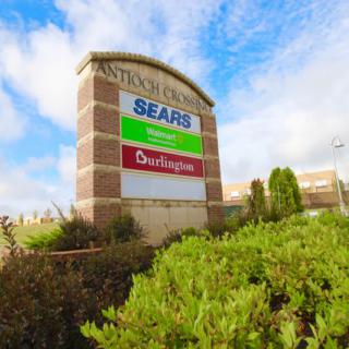 A shopping mall sign displaying storefronts like Sears and AutoNation against a partly cloudy sky.