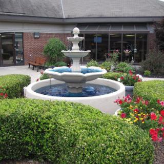 A serene garden scene featuring a multi-tiered fountain surrounded by vibrant flowers and neatly trimmed bushes.