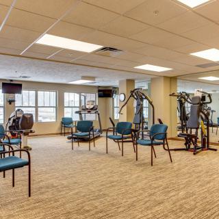 A spacious gym with exercise equipment and chairs, featuring large windows and a mirrored wall.