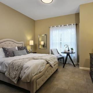 A cozy bedroom with a neatly made bed, nightstands with lamps, and a small seating area by the window.