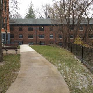 A view of a brick apartment building with a concrete pathway lined by grass and trees in winter.