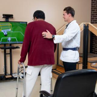 A physical therapist assists a patient using a walker during a rehabilitation exercise with a video game.