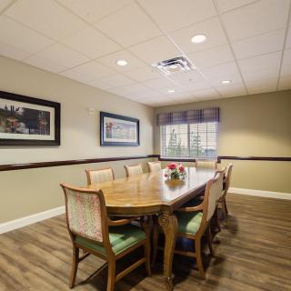 A well-lit dining or meeting room with a long wooden table and chairs, framed artworks on the walls, and a large window.