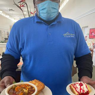 A person in a blue shirt holding two plates with food and dessert, inside a restaurant.