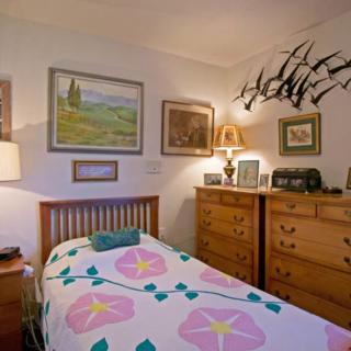 A cozy bedroom with a single bed adorned with a floral quilt, surrounded by artwork and wooden furniture.