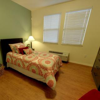 A cozy bedroom with a single bed, decorated with patterned bedding and cushions. The room features wooden flooring, a bedside lamp, and window blinds.