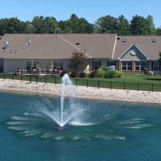 A scenic view of a water fountain in a pond, surrounded by a residential building and lush greenery.
