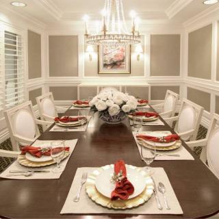 An elegant dining room with a beautifully set table featuring white and gold decor, red napkins, and a centerpiece of white flowers under a chandelier.