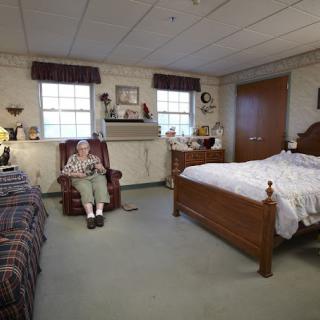 An elderly person sitting in an armchair in a cozy bedroom with a bed and decorative items.