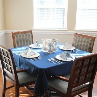 A dining table with a blue tablecloth and place settings for four in a sunlit room.