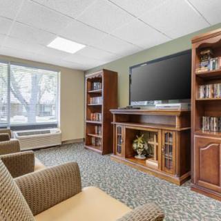 A cozy lounge area featuring comfortable chairs, a large television with wooden bookshelves, and a window letting in natural light.