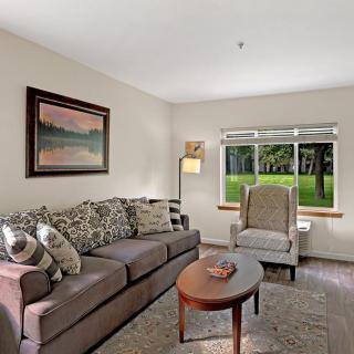 A cozy living room with a sofa, armchair, and a coffee table, featuring a large window and a painting on the wall.