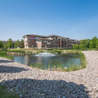 A modern building overlooks a serene pond with a fountain, surrounded by rocks and lush greenery on a sunny day.