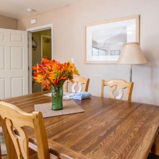 A cozy dining room featuring a wooden table adorned with a vase of vibrant orange flowers, surrounded by matching chairs and complemented by soft lighting and wall art.