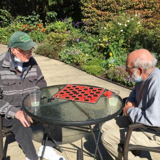 Two elderly men play a game of checkers outdoors in a garden setting on a sunny day.